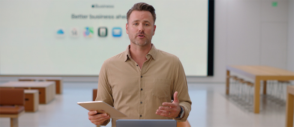 Moderator spricht und hält dabei ein iPad in der Hand, vor ihm steht ein MacBook auf einem Tisch, während ein großes Display hinter ihm verschiedene Business App Symbole anzeigt.