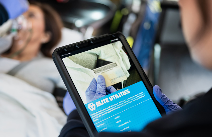 The Elite Utilities app is open on iPad and a paramedic scans a patient’s drivers licence while the patient lays on a transport bed