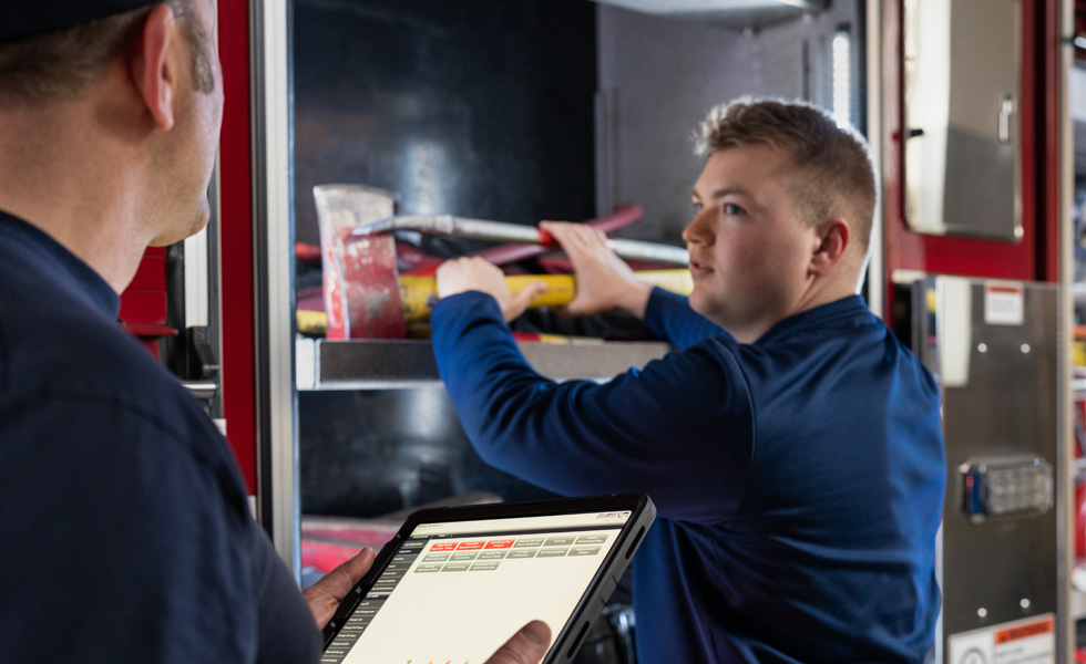 Two crew members inspect tools and equipment on a fire engine, one holds an iPad while the other inspects 