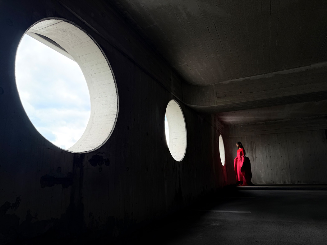 A woman wearing a long red dress stands in an empty room being photographed. The Photo app toggles through the following focal lengths to demonstrate the capabilities of the pro camera system: 200 mm, 100 mm, 48 mm, 35 mm, 28 mm, 13 mm, and macro