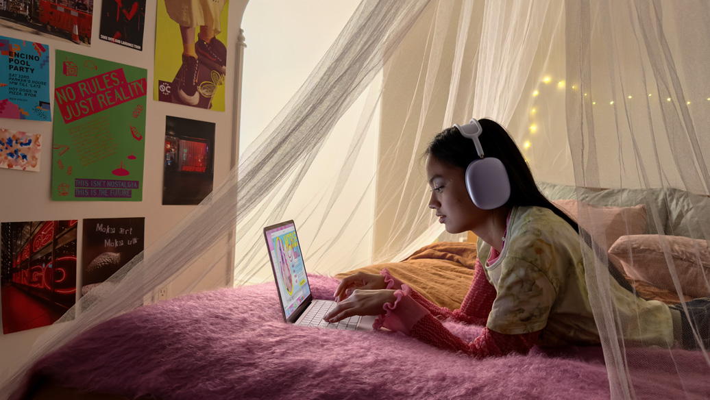 A young person in their bedroom wearing headphones uses MacBook Neo, blush colour, to book a nail appointment