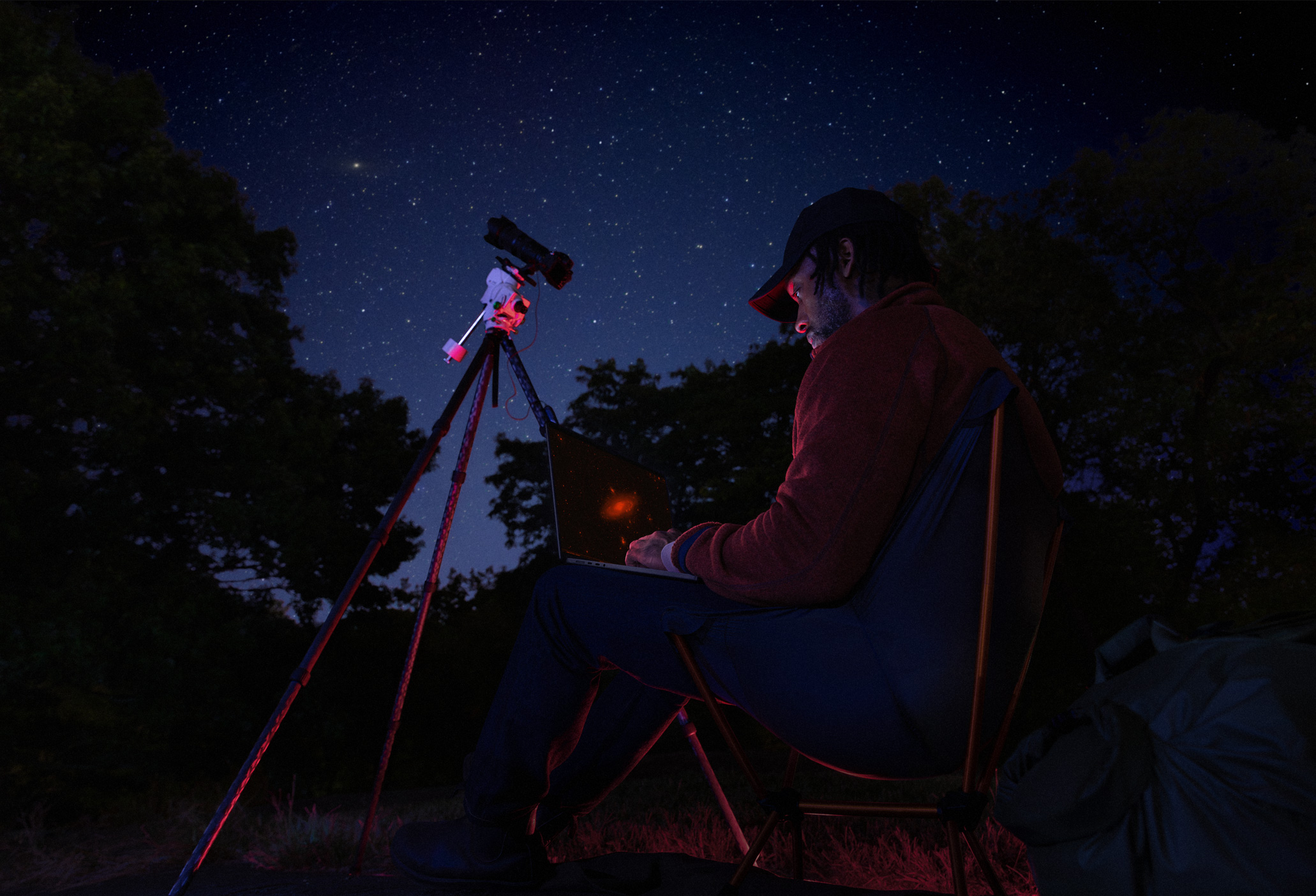 A person outside at night using a MacBook Pro on their lap, connected to a device on a tripod pointed towards the starry sky
