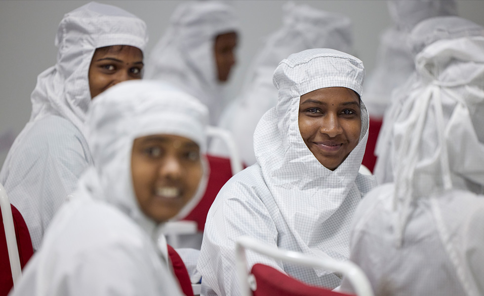 Several employees sit in chairs with full-coverage work suits, but three are turned towards the camera and smiling