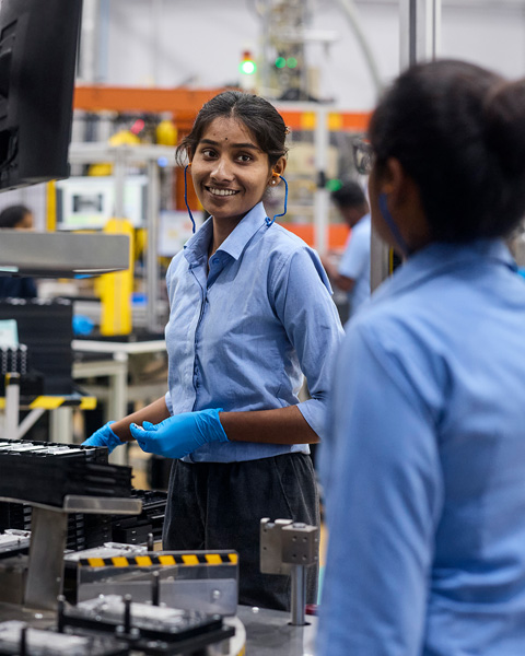 An employee wearing protective earbuds in a component manufacturing facility smiles at a co-worker