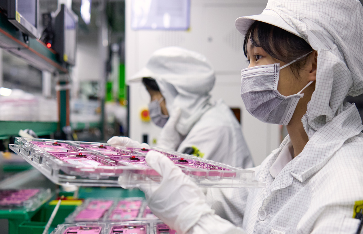 Two employees wearing full-coverage work suits handle iPhone parts in an assembly facility