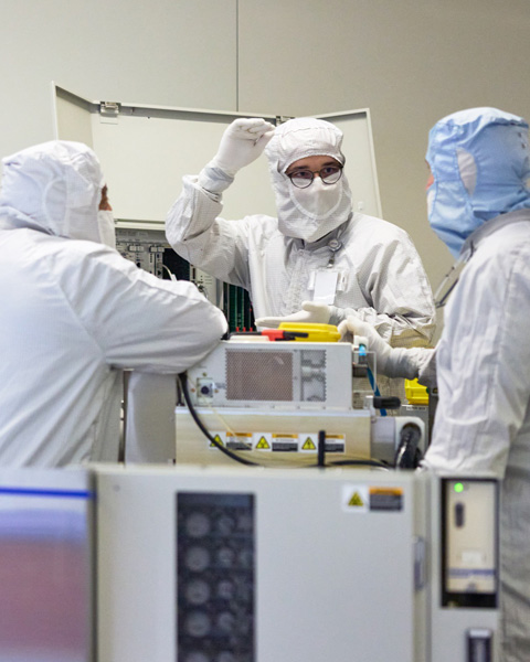 Three employees in full-body work suits stand and collaborate around a silicon manufacturing machine