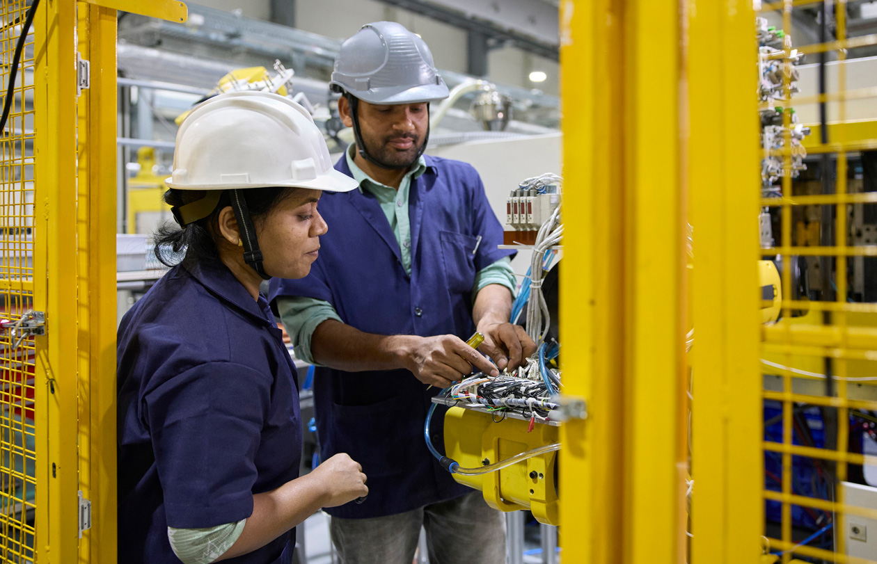 Twee medewerkers in een werkoverall en met een veiligheidshelm staan bij hun werkplek en bedienen assemblage-machines