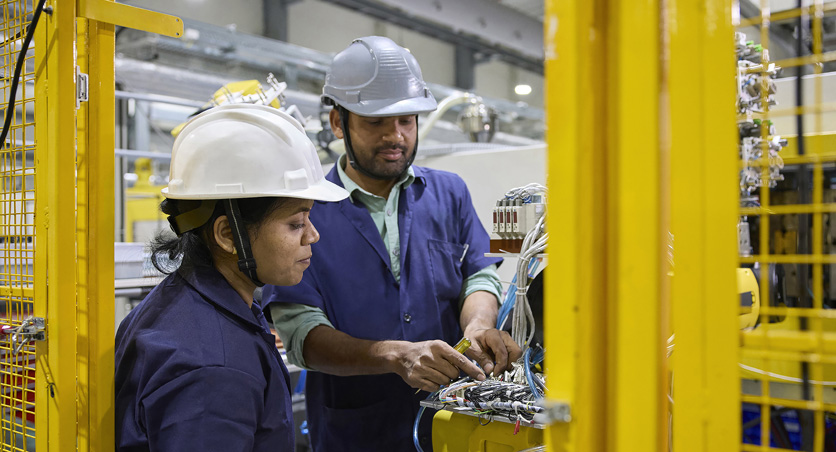 Twee medewerkers in werkoveralls en met veiligheidshelmen staan voor een assemblage-machine